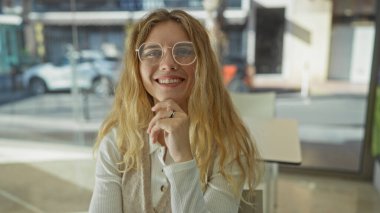 Ofiste sarışın ve gözlüklü bir kadın gülümsüyor. Parlak bir günde pencereden gün ışığının tadını çıkarıyor. Olumlu ve profesyonel bir tavır sergiliyor..