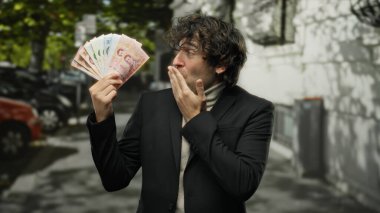 Şehir caddesinde elinde şaşırmış bir ifadeyle Thai banknotları tutan, resmi takım elbiseli, İspanyol kökenli, bulanık şehir geçmişli gün ışığı, paraya odaklanmış bir adam.