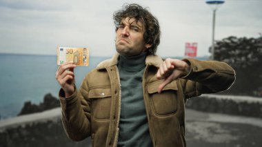 Hispanik bir adam sahilde elinde avroluk banknotla okyanus zemininde memnuniyetsizliğini dile getiriyor..
