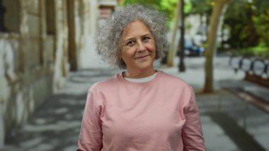 Kentteki ağaçlar ve binalarla dolu bir sokakta duran pembe kazaklı, kendine güvenen gri saçlı bir kadın..