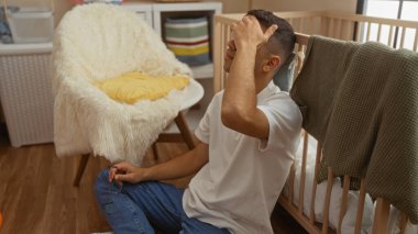 Kapalı bir odada, bebek beşiğinin yanında oturan genç adam. Gözlüklü ve yanında rahat bir sandalyeyle düşünceli görünüyor.