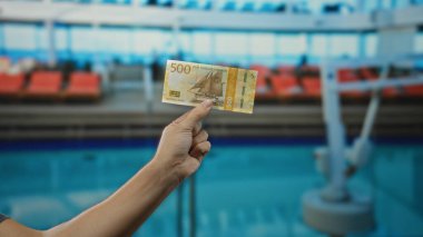 Norveçli bir adam otelin açık havuz kenarında banknot tutuyor, mavi su arkaplanı ve parlak tatil atmosferiyle boş bir ortamda para gösteriyor..