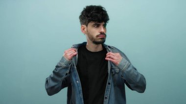 Genç adam kot ceketini mavi arka plana göre ayarlıyor, şık ve düşünceli görünüyor, modern moda ve kendine güveni bir nebze de olsa gösteriyor..