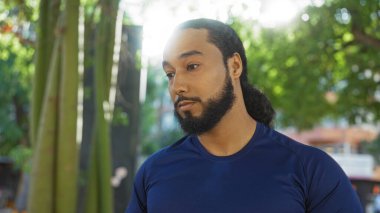 Şehir parkında sakallı genç bir adam gündüz vakti şehir yeşillikleri arasında çeşitli kültürleri ve dingin ifadeyi yansıtıyor. Modern yaşam tarzını açık havada sergiliyor.