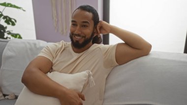 Sakallı genç adam rahat oturma odasında koltukta dinleniyor, ev atmosferinin tadını çıkarırken gülümsüyor ve rahat yastığa yaslanıyor modern apartman dekoruyla birlikte..