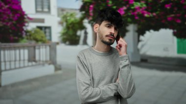 Siyah saçlı ve gri kazaklı genç adam arka planda çiçekleri bulanık bir şekilde şehir sokağında akıllı telefondan konuşuyor.