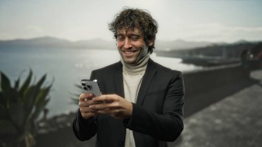 Sahilde akıllı telefonları kullanarak gülümseyen genç adam, sakin bir sahil kenarında modern yaşam tarzını ve bağlantılarını yakalıyor..