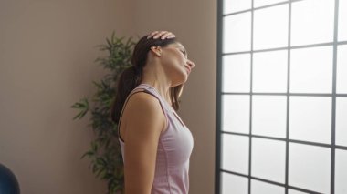 Spor salonunda boynu büyük camlı, esmer kadın içeride spor kıyafetleri giyiyor, içeri ışık giriyor, sakin atmosfer, spor yaparken odaklanmış ifade..