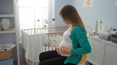 Bebek beşiğinin yanında rahat bir yatak odasında oturan hamile bir kadın, şefkatle karnını tutuyor, besleyici bir beklenti içinde, rahatlatıcı bir dekor ile çevrili, annelik atmosferini geliştiriyor..