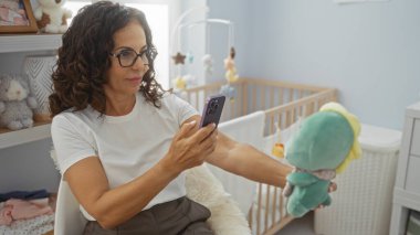 Kadın, rahat bir yatak odasında beşiği olan, yumuşak oyuncaklarla çevrili bir şekilde rahat bir şekilde otururken telefonla anları yakalıyor..