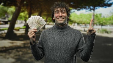 Parkta gülümseyen İspanyol bir adam parmaklarıyla yukarıya doğru işaret ederek doğal bir ortamda başarı, zenginlik ve iyimserliği temsil ediyor..