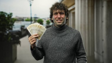 Hispanik bir adam Norveçli krone banknotlarını elinde tutarken kendine güveni tam bir tavır sergiliyor, finansal başarıyı ve kentsel yaşam tarzını vurguluyor..