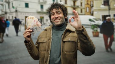 İspanyol adam elinde Türk lirası tutarak hareketli bir şehir caddesinde güvenle gülümsüyor, canlı şehir hayatının ortasında bir onay jesti ile paralarını sergiliyor..