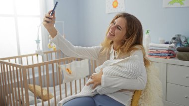 Yeni doğmuş bebeğiyle sıcak bir yatak odasında selfie çekerken gülümseyen bir kadın, anneliği neşeyle kucaklıyor ve güzel anları kapalı bir yerde sevgiyle yakalıyor..