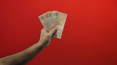 İzole edilmiş kırmızı arkaplan karşısında Türk lira banknotları tutan adam, mali temalara vurgu yapıyor ve Türk para birimini canlı bir ortamda vurguluyor.