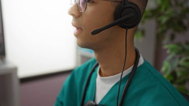 Hastane odasında steteskoplu kulaklık takan genç adam klinik ortamlarda çalışırken odaklanmış görünüyor ve hastalara telefonla yardım ediyor..