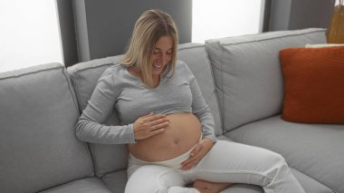 Kadın rahat bir oturma odasında rahat bir şekilde karnına dokunurken yumuşak bir kanepede oturup annelik beklentisini ve neşesini dile getiriyor.