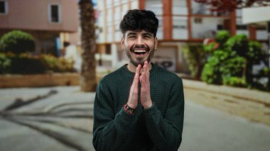 Neşeli bir suratı olan genç adam güneşli bir günde ağaçların ve binaların olduğu bir şehirde eğleniyor..