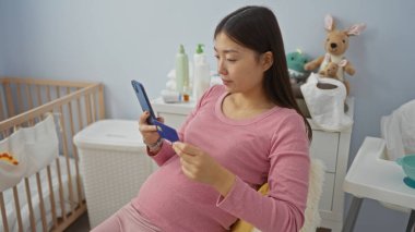 Hamile kadın yatak odasında bebek beşiği ve oyuncaklarıyla telefon ve kart kullanıyor. Evde rahat ve annelik atmosferi yaratıyor. Modern annelik detaylarını vurguluyor..