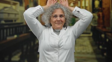 Gri saçlı yaşlı bir kadın kilisede neşeli jestler yapıyor, beyaz elbiseler ve gözlük takıyor, kutsal ortama mizah ve kapris katıyor..