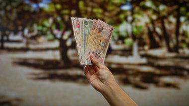 Güneşli bir şehir parkı ortamında elinde Dirham banknotları tutan adam, şehir arka planının bulanık olduğu doğal bir sokak ortamında para gösteriyor..
