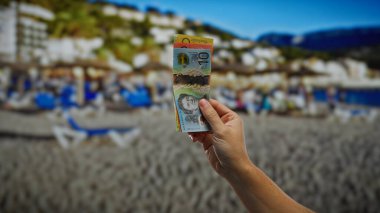 Denizin kenarında güneşli bir sahilde el ele tutuşmuş, şehir arkaplanı bulanık, açık deniz kenarında finansal bir konsepti tasvir ediyor..