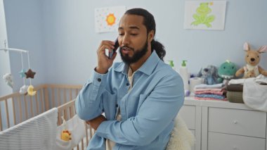 Bebek odasında beşikte ve oyuncaklarla telefonda konuşan sakallı genç adam, babalık ve aile hayatını ima ediyor, mavi giyinmiş ve yumuşak renklerle çevrili..