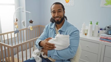 Açık mavi duvarları, beşiği ve bebek ihtiyaçlarıyla bebeği kucağında tutan gülümseyen bir adam. Bakımını ve huzur dolu ev ortamını yansıtıyor..