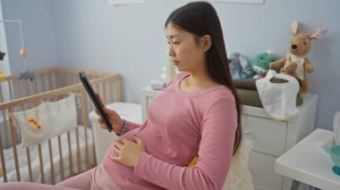 Bebek odasında beşikteki oyuncaklarla ve modern dekorla tablet kullanan hamile bir kadın, sakinliğini ve aile hazırlama ruh halini evin içinde aktarıyor.