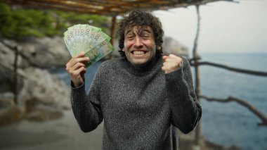 İspanyol adam, deniz kıyısında rusya rublesini neşeyle tutuyor, açık havada süveter içinde heyecanını ifade ediyor..