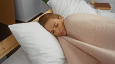 Sıcak bir yatak odasında beyaz yastıklı bir yatakta uzanan, yumuşak bir battaniyeye sarılmış, modern bir ev ortamında huzurlu bir ev ortamını sergiliyor..