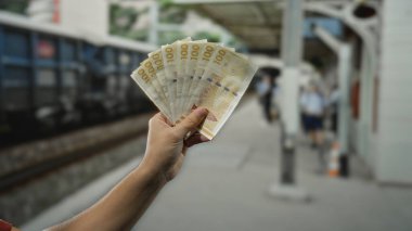 Bir açık hava tren istasyonunda Danimarka kronu banknotları tutan bir adam, halka açık bir demiryolu ortamında yerel para birimini gösteriyor, seyahat ve finansal kavramları vurguluyor.