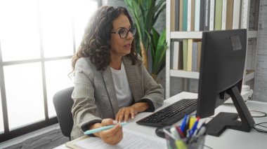 Ofiste çalışan İspanyol bir kadın, bilgisayar görevlerine odaklanmış, içi kitaplarla ve büyük pencerelerle dolu modern bir iç mekan ile çevrili, profesyonelliği ve konsantrasyonu aktarıyor..