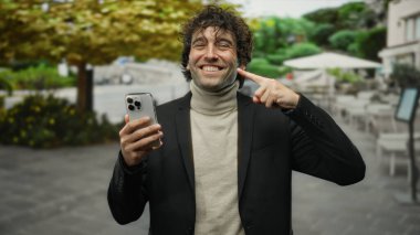 Genç İspanyol bir adam parmaklarıyla dişlerini göstererek gülümsüyor ve bir sokakta telefon tutuyor, şehir ortamında kendine güveni ve tarzı var..
