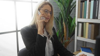 Modern ofiste kitaplıklar ve bitkilerle çalışan bir kadın profesyonel ve odaklanmış bir atmosfer sergileyerek telefonla konuşuyor..