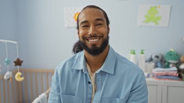 Sakallı, gülümseyen bir adam, bebek beşiği ve eğlenceli dekoruyla rahat bir yatak odasında oturur ve evin içinde sıcak ve davetkar bir atmosfer yaratır..