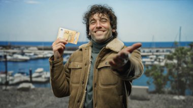 Deniz kenarında gülümseyen İspanyol bir adam, elinde marinası ve arka planında tekneleri olan 50 avroluk banknotla elini kameraya doğru uzatıyor..