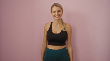 İzole edilmiş pembe arka planda gülümseyen genç bir kadın spor kıyafetleri giyerek pozitif enerji ve zindelik yaşam tarzı gösteriyor..