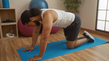 Sakallı genç adam sağlık merkezinde mavi minder üzerinde egzersiz yapıyor. Etrafında spor aletleri var. İçeride sakin ve odaklanmış bir çalışma ortamı sunuyor..