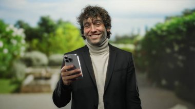 Parkta gülümseyen genç bir adam elinde akıllı bir telefonla, doğal yeşil bir arka planla neşe ve bağlantıyı ifade ederek sakin ve dostane bir atmosfer yaratıyor..