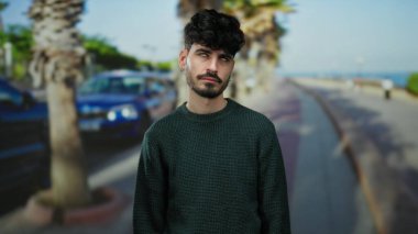 Arka planda palmiye ağaçları olan kıvırcık saçlı yakışıklı genç adam rahat ama düşünceli bir tavır sergiliyor..