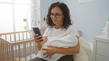 Bir kadın yatak odasında, battaniyeye sarılı bebeği tutarken akıllı telefona bakıyor ve evde sakin bir aile anı yaşıyor..