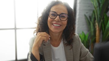 Ofis masasında gülümseyen olgun bir kadın doğal ışıkla arkadaşça ifadesini ve mesleki davranışını vurguluyor..