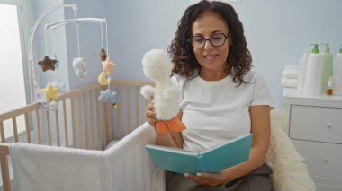 Yatak odasındaki bir sandalyede oturan bir kadın elinde oyuncak ve bir kitapla bebeğin beşiğinin yanında, besleyici, rahatlatıcı ve eğitici bir ortam sunuyor..