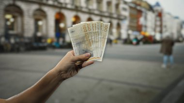 Bir şehir sokak ortamında Danimarka kronu banknotları el ele tutuşmuş, kentsel arkaplan karşısında finansal değişimi vurguluyor, insanın figürü ve tarihi mimarisi görünür..