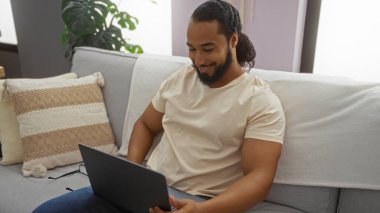 Modern oturma odasında dizüstü bilgisayar kullanan sakallı genç adam doğal ışık ve rahat oturma düzeniyle rahat bir iç mekan sergiliyor.