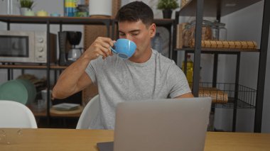 Laptop 'ta çalışırken kahve içen genç adam, raflar, mikrodalga fırın ve diğer ofis eşyalarıyla çevrili, modern bir iş yeri ortamını yansıtıyor..