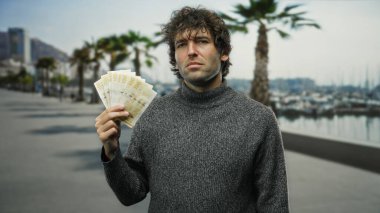 Norveç kronu taşıyan adam deniz kıyısındaki bir limanda, arka planda botlar ve palmiye ağaçları ile birlikte, seyahat ve finans duygusunu aktarıyor..