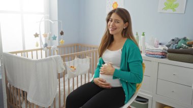 Bebek eşyalarıyla çevrili, rahat bir bebek odasında otururken gülümseyen hamile bir kadın, parlak bir ev ortamında karnını mutluluk ve beklentiyle kucaklıyor..
