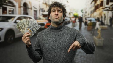 İspanyol asıllı bir adam şehir sokaklarında Amerikan dolarlarını tutuyor, şehir içi dolandırıcılıkla çevrili, açık bir ortamda kendine güvenen bir ifadeyle zenginliği vurguluyor..